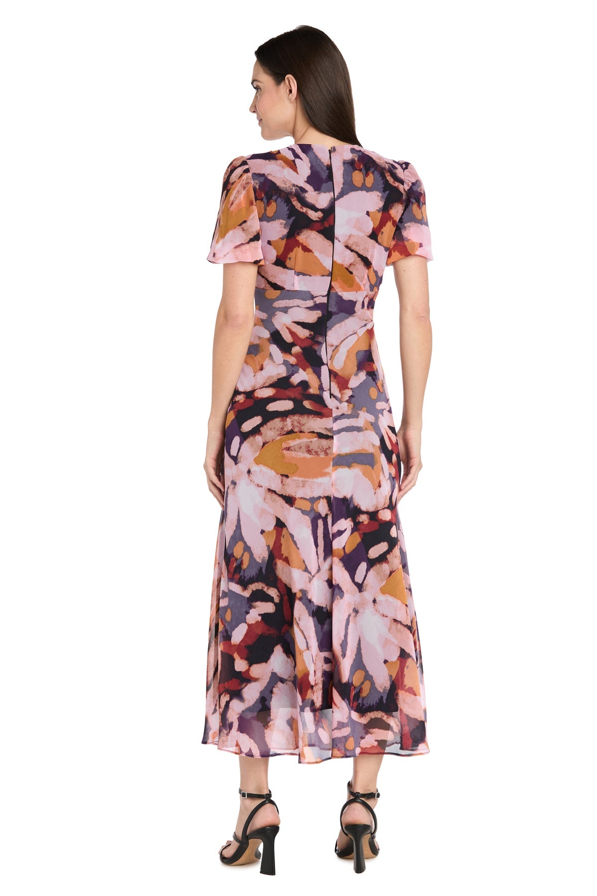 A woman faces away wearing the R&M Richards 4161 short sleeve print party dress in a colorful abstract pattern, paired with black heeled sandals—an ideal choice for a wedding guest outfit.